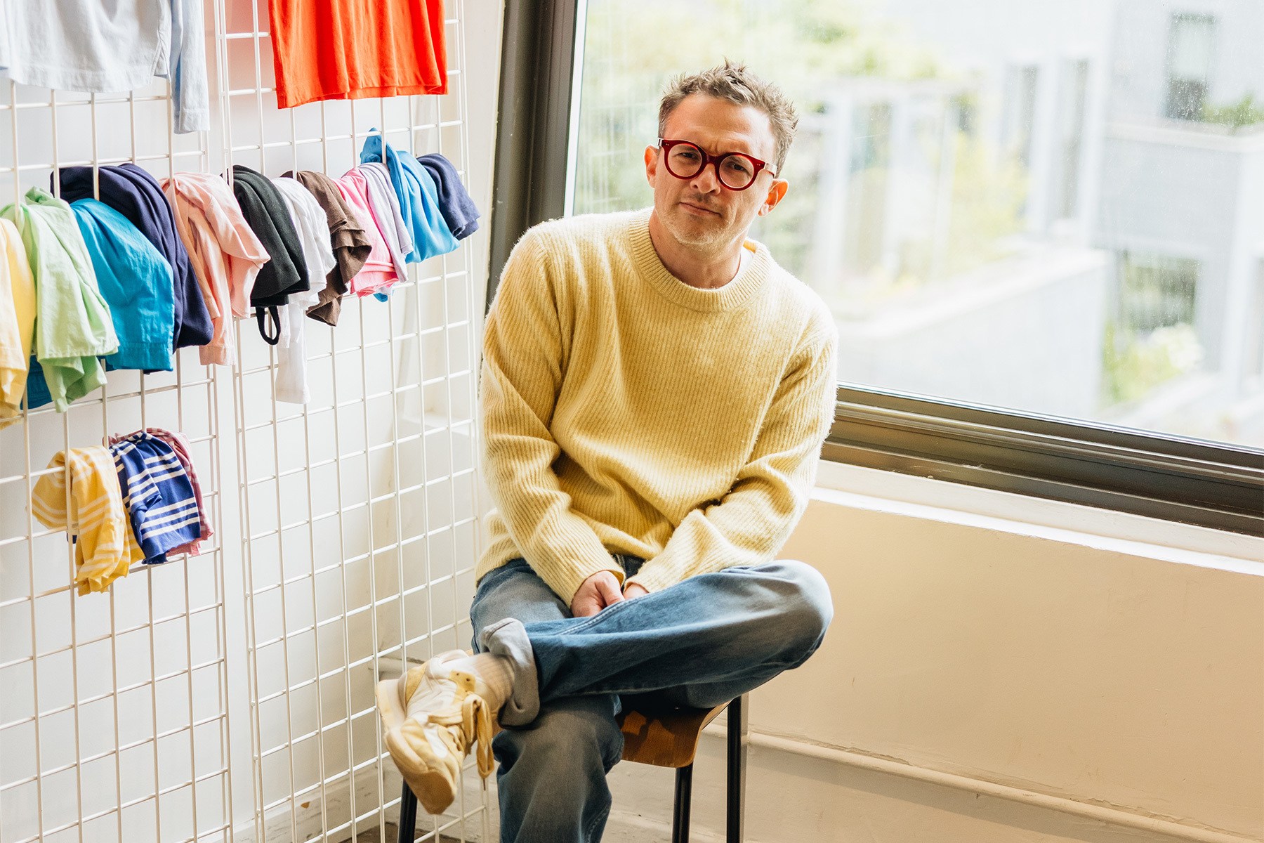 Man in yellow sweater and red glasses sits on a stool, with colorful clothes hanging on a grid behind him.
