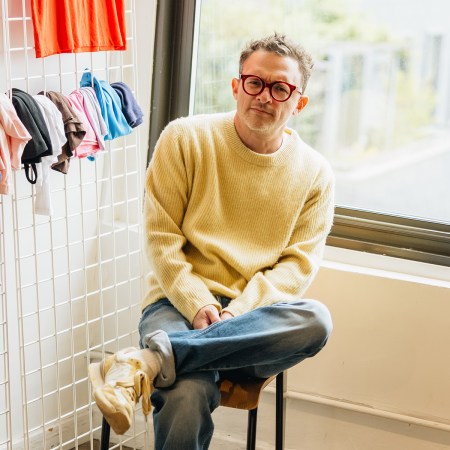 Man in yellow sweater and red glasses sits on a stool, with colorful clothes hanging on a grid behind him.
