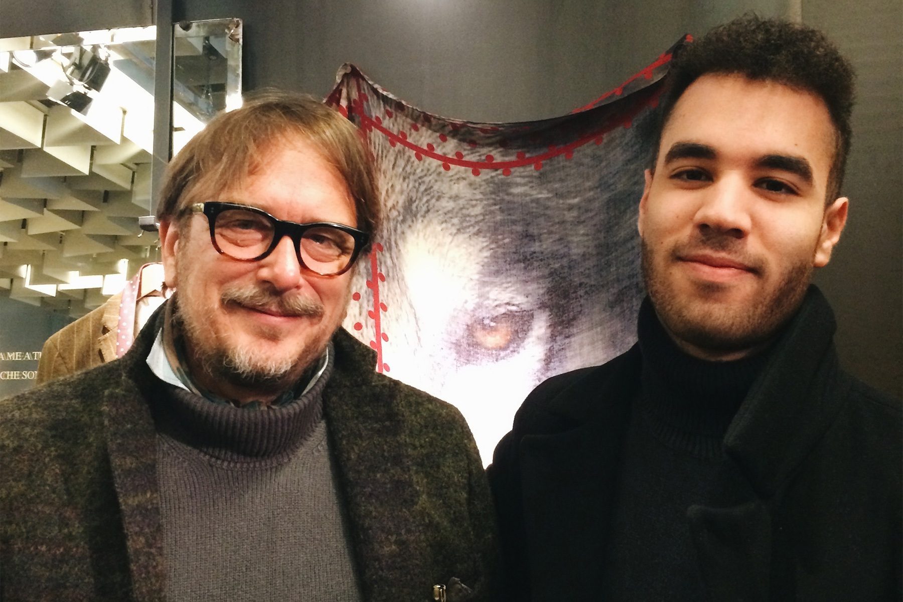 Two men, one older with glasses and a mustache, one younger with a beard, stand smiling in front of a wolf poster.