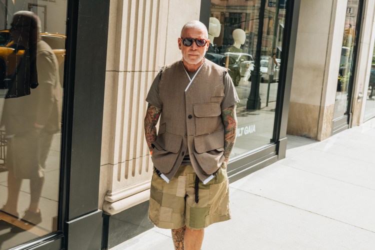 A man with a shaved head, sunglasses, and heavily tattooed arms wears a deconstructed brown vest over a gray t-shirt and patchwork shorts.