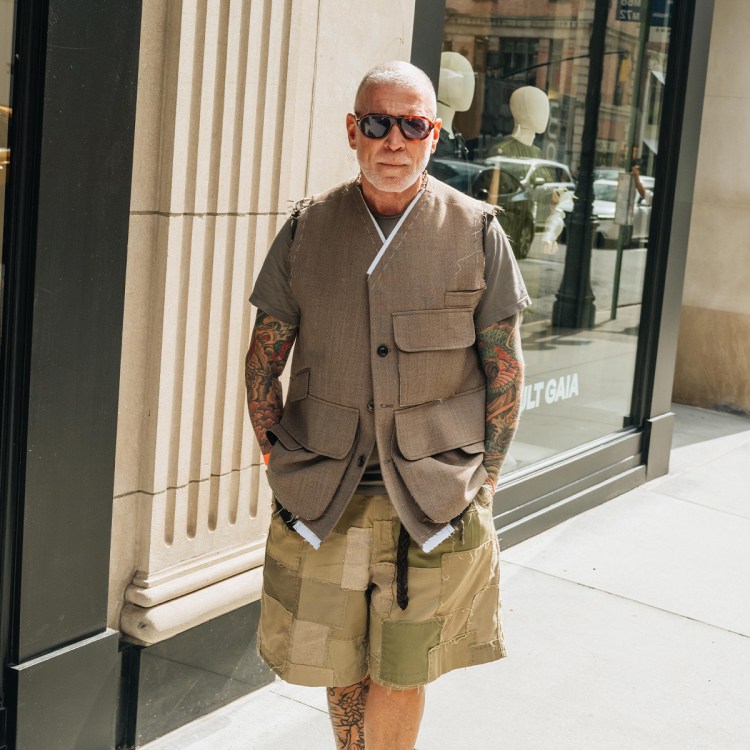 A man with a shaved head, sunglasses, and heavily tattooed arms wears a deconstructed brown vest over a gray t-shirt and patchwork shorts.