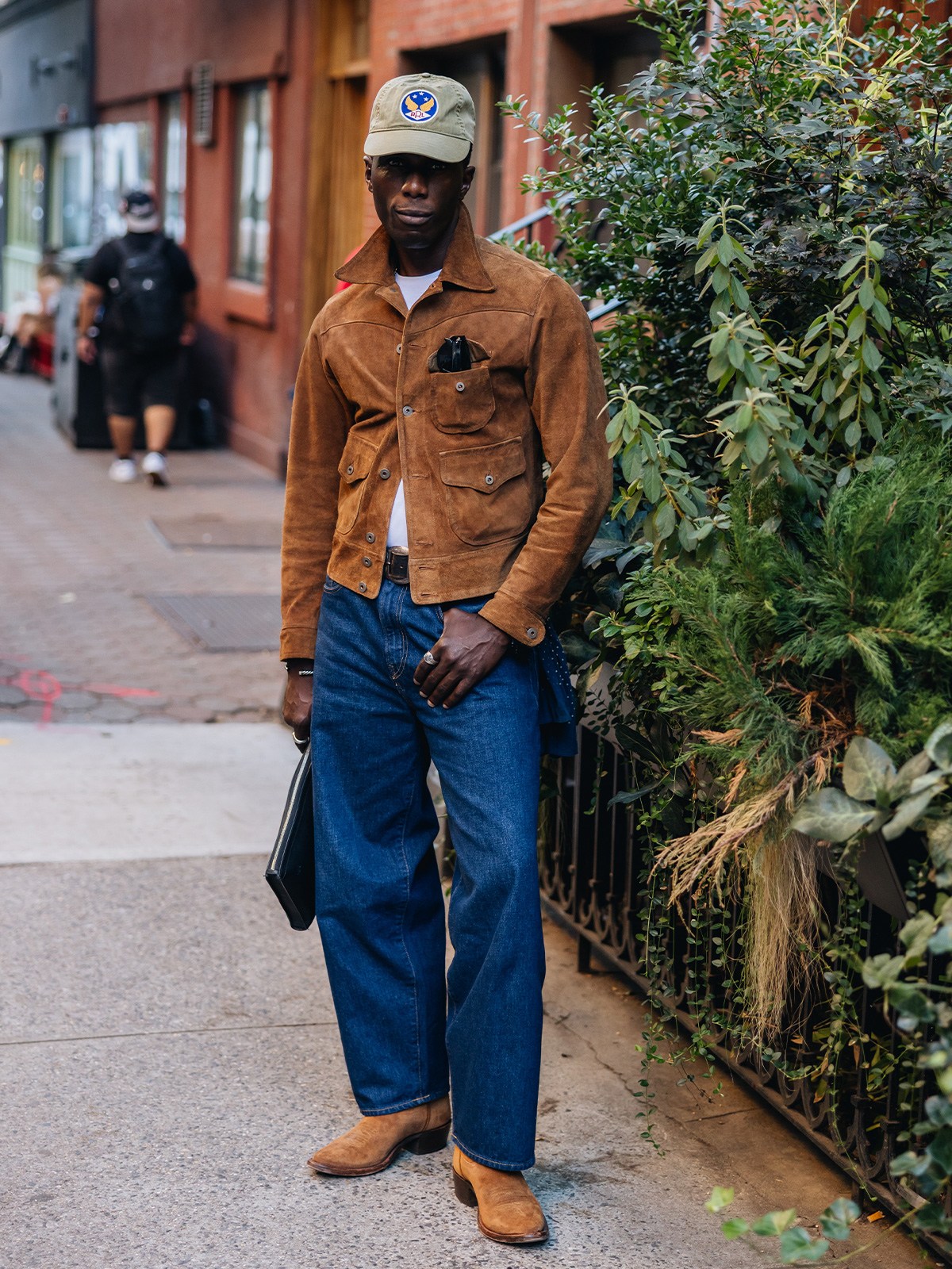 A man in a brown suede jacket, wide-leg jeans, and cowboy boots stands on a city sidewalk.