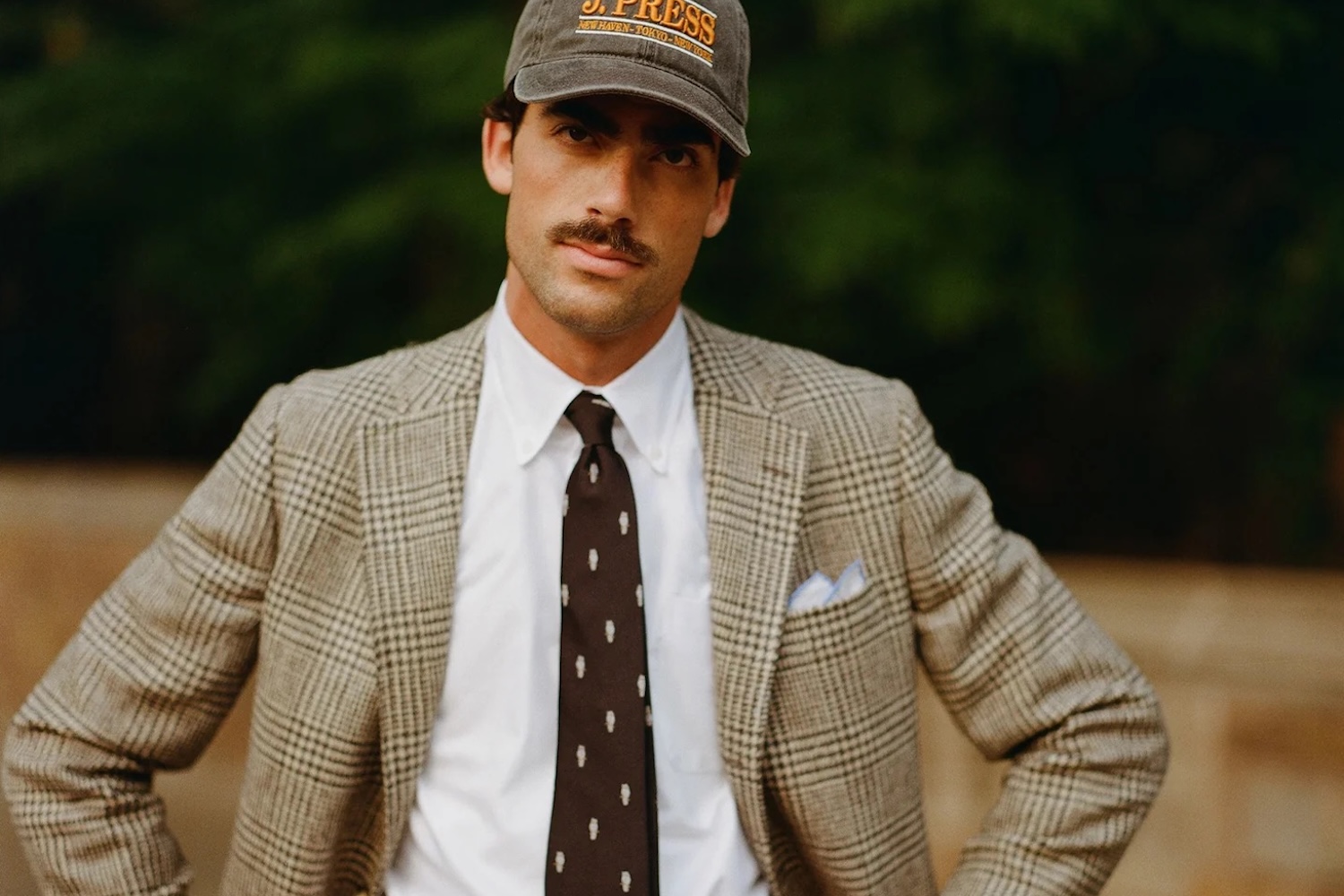 Man with a mustache wearing a plaid blazer, white shirt, dark tie with white figures, and a J. Press baseball cap.