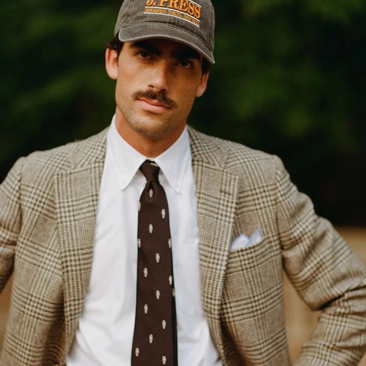 Man with a mustache wearing a plaid blazer, white shirt, dark tie with white figures, and a J. Press baseball cap.