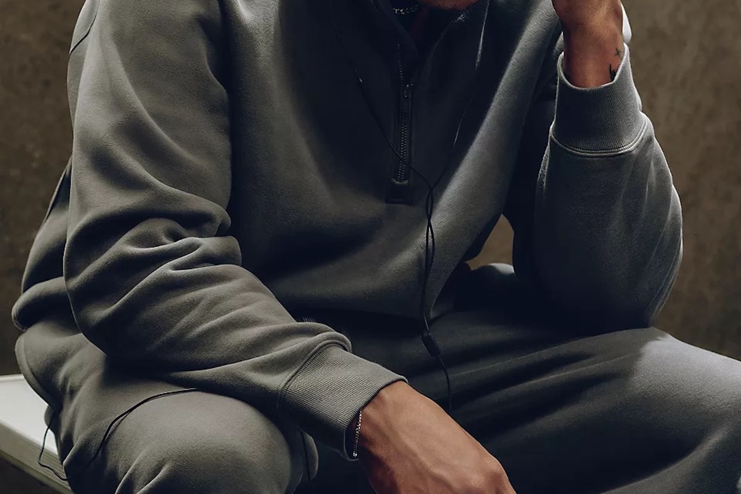 Person in a gray sweatsuit with a half-zip top and wired earbuds, resting their arm on their knee.