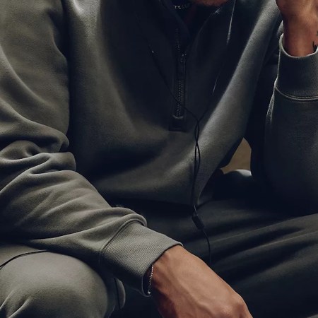 Person in a gray sweatsuit with a half-zip top and wired earbuds, resting their arm on their knee.