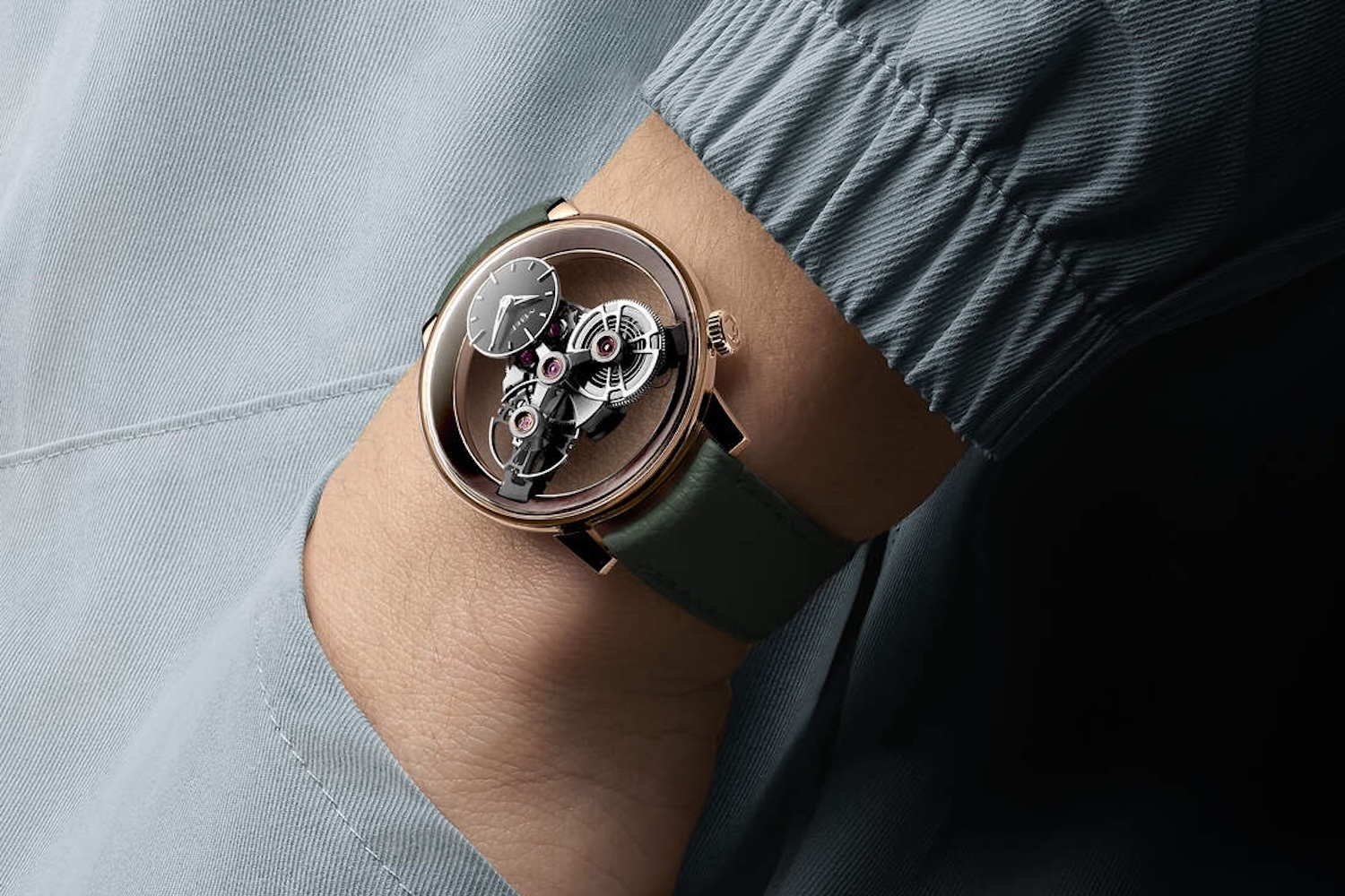 A person's arm wearing a rose gold watch with a clear face revealing intricate gears, on a dark green leather strap.