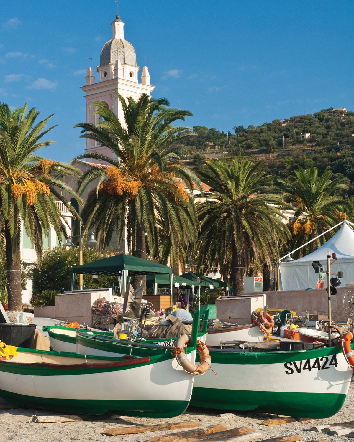 Green and white fishing boats on a sandy beach, with palm trees, a pink church tower, and a green hillside in the background.