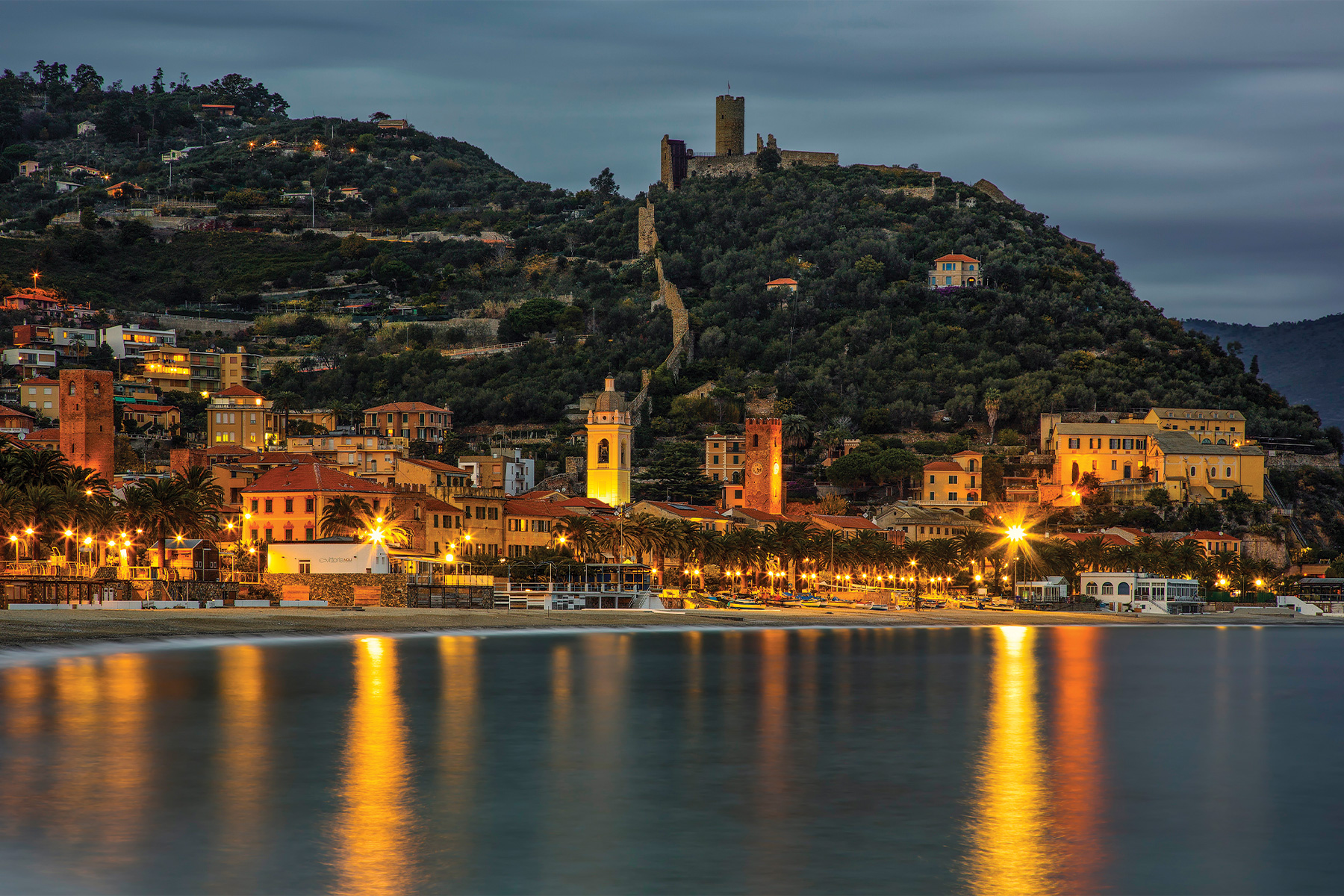 Coastal town at dusk with illuminated buildings, palm trees, and a beach, reflecting lights on the calm water.