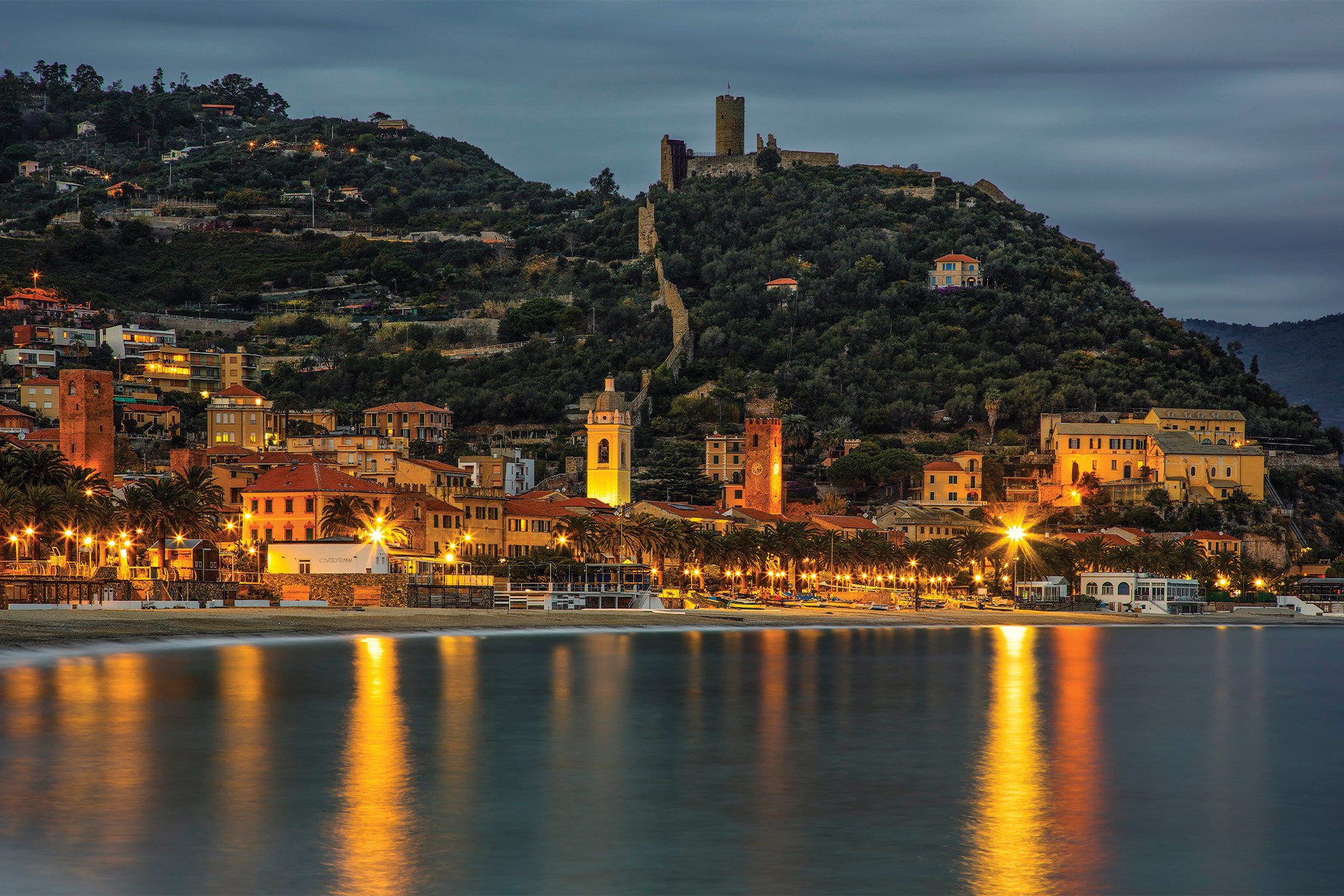 Coastal town at dusk with illuminated buildings, palm trees, and a beach, reflecting lights on the calm water.