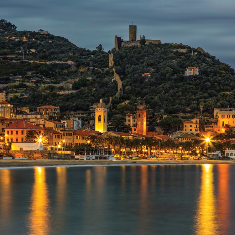 Coastal town at dusk with illuminated buildings, palm trees, and a beach, reflecting lights on the calm water.