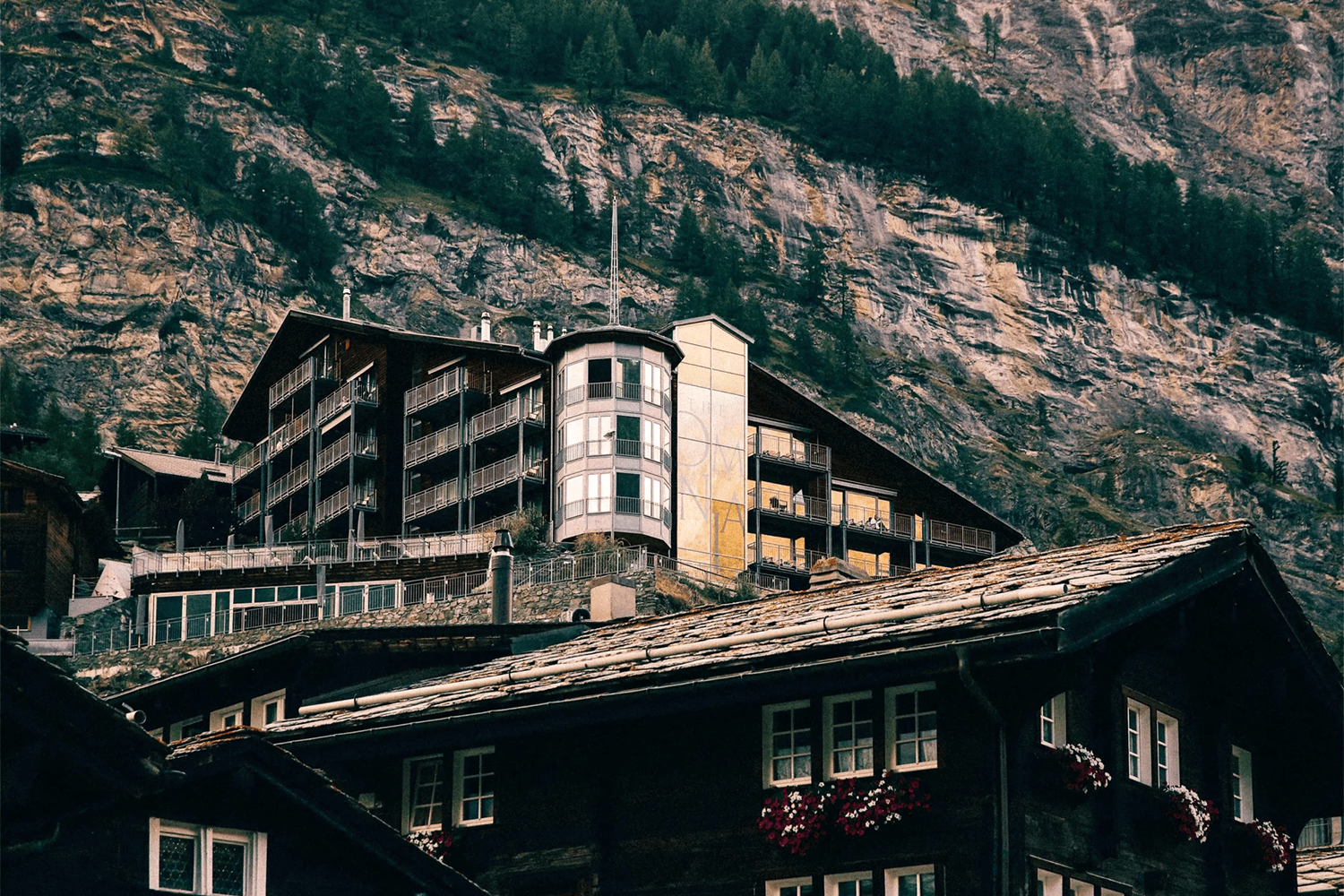 Multi-story buildings with balconies on a steep, rocky mountainside, with a dark wooden building in the foreground.