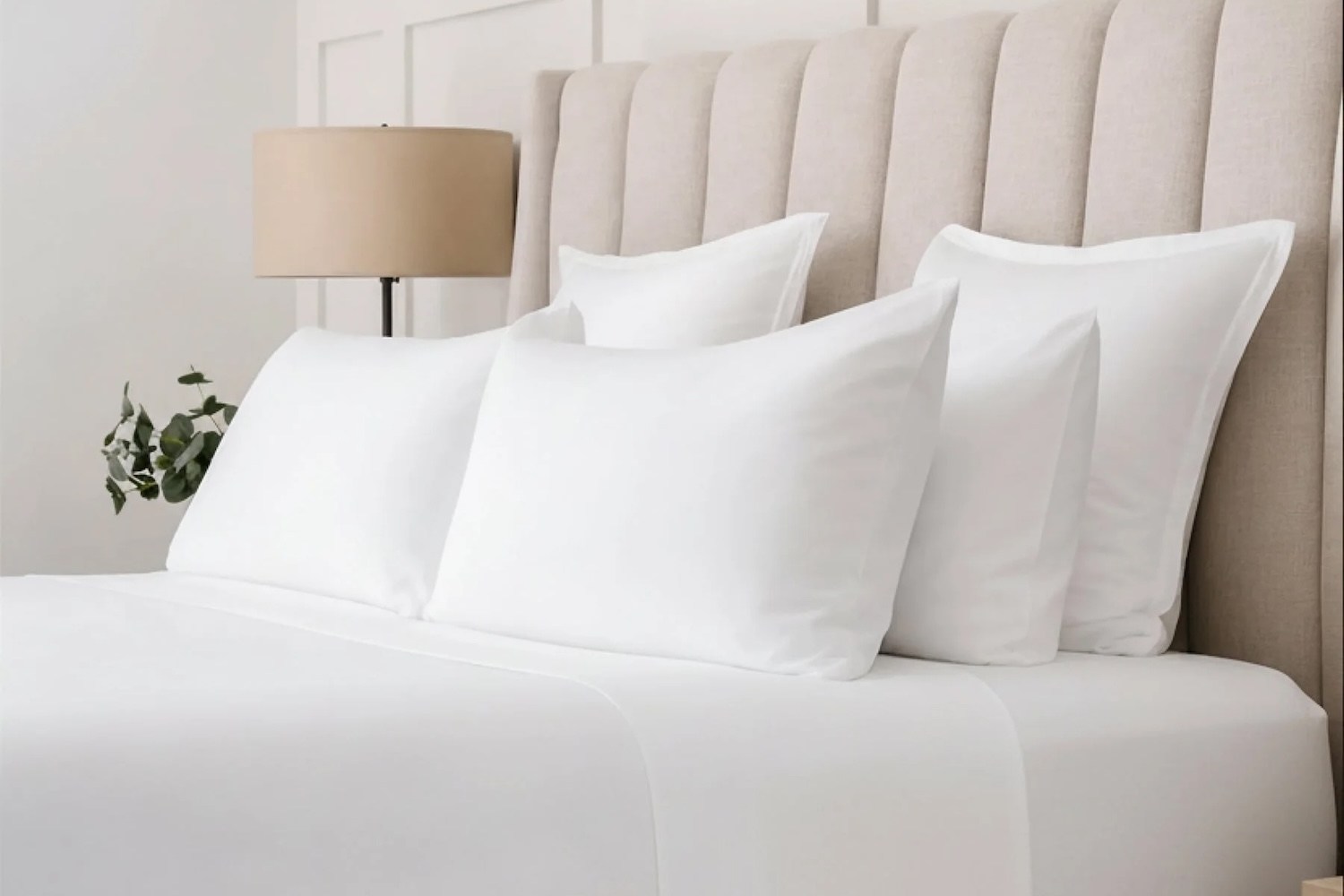 A bed with white sheets and four white pillows, a beige upholstered headboard, a lamp, and a plant.