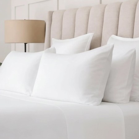 A bed with white sheets and four white pillows, a beige upholstered headboard, a lamp, and a plant.