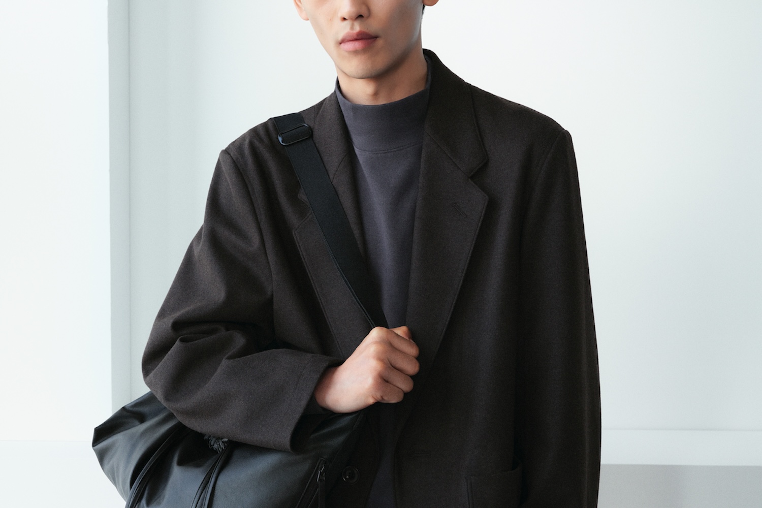 A person in a dark brown blazer and gray turtleneck holds a black messenger bag.