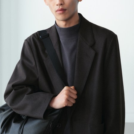 A person in a dark brown blazer and gray turtleneck holds a black messenger bag.
