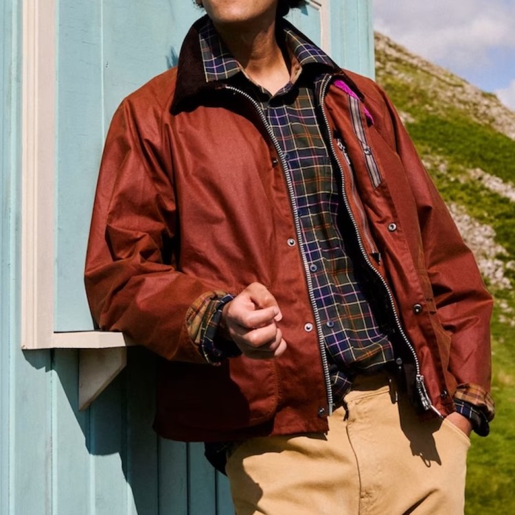 Person in a brown waxed jacket, plaid shirt, and tan pants leaning on a light blue wall, with a green hillside in the background.