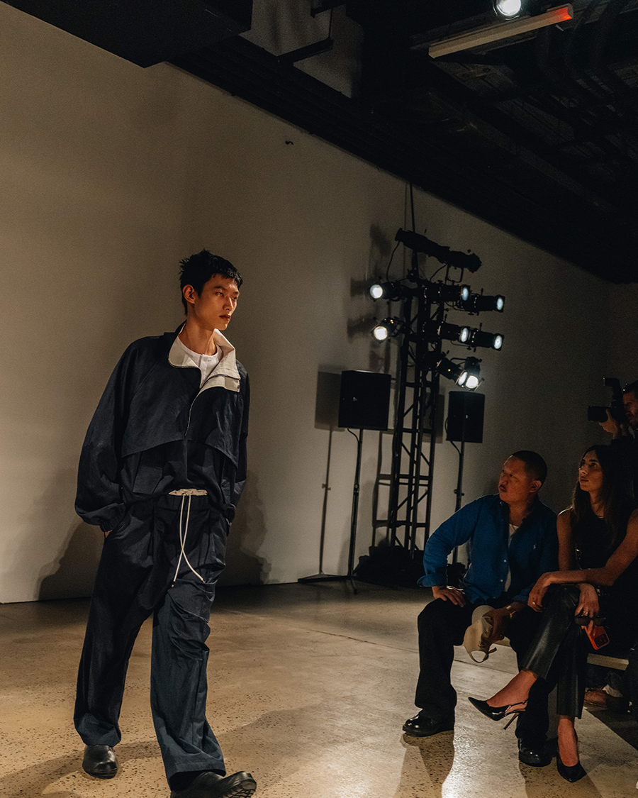 A male model in a dark blue tracksuit walks a runway, with audience members and stage lights in the background.