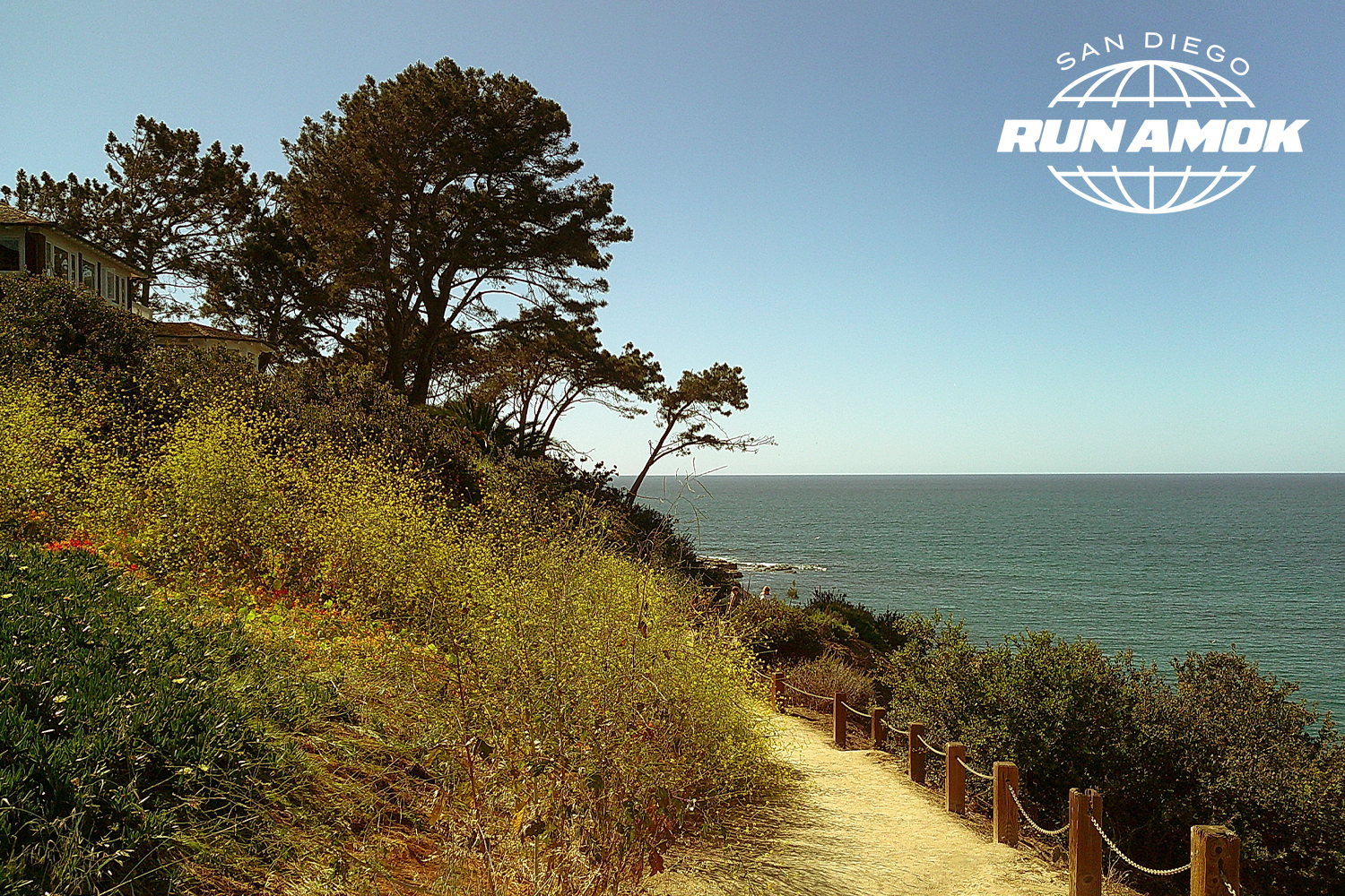 Dirt path with a rope fence leading along a cliffside with green and yellow foliage, overlooking a calm blue ocean.