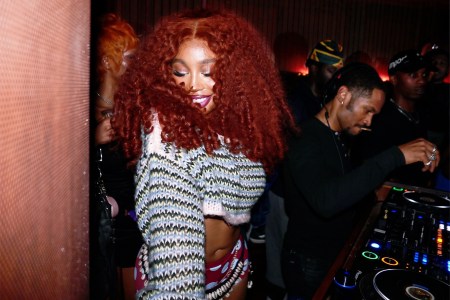 SZA, with voluminous red curly hair and a cropped sweater, smiles while a DJ works the turntables in a dimly lit club.
