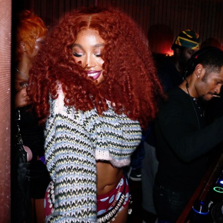 SZA, with voluminous red curly hair and a cropped sweater, smiles while a DJ works the turntables in a dimly lit club.