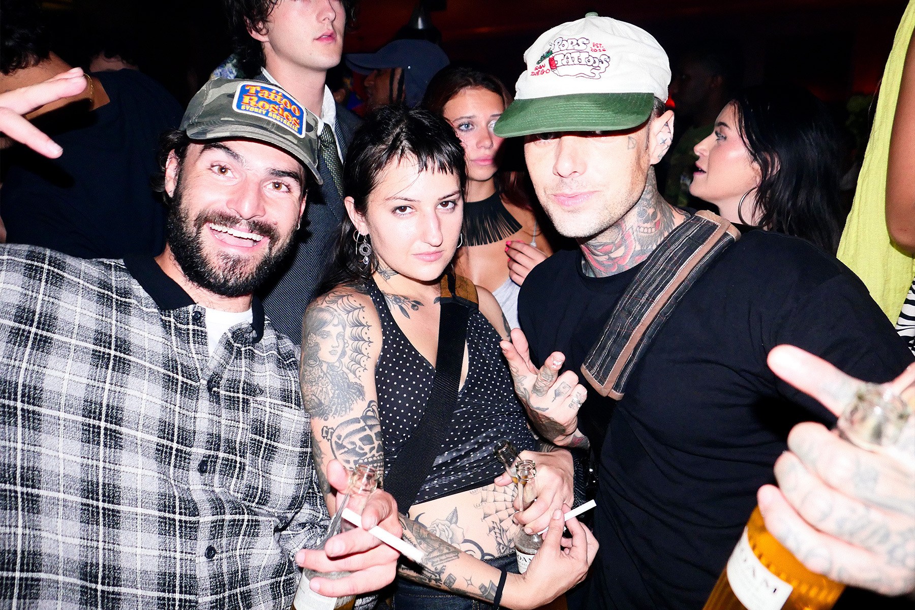 Three tattooed people, two men and one woman, pose for a photo at a party, holding drinks and cigarettes.