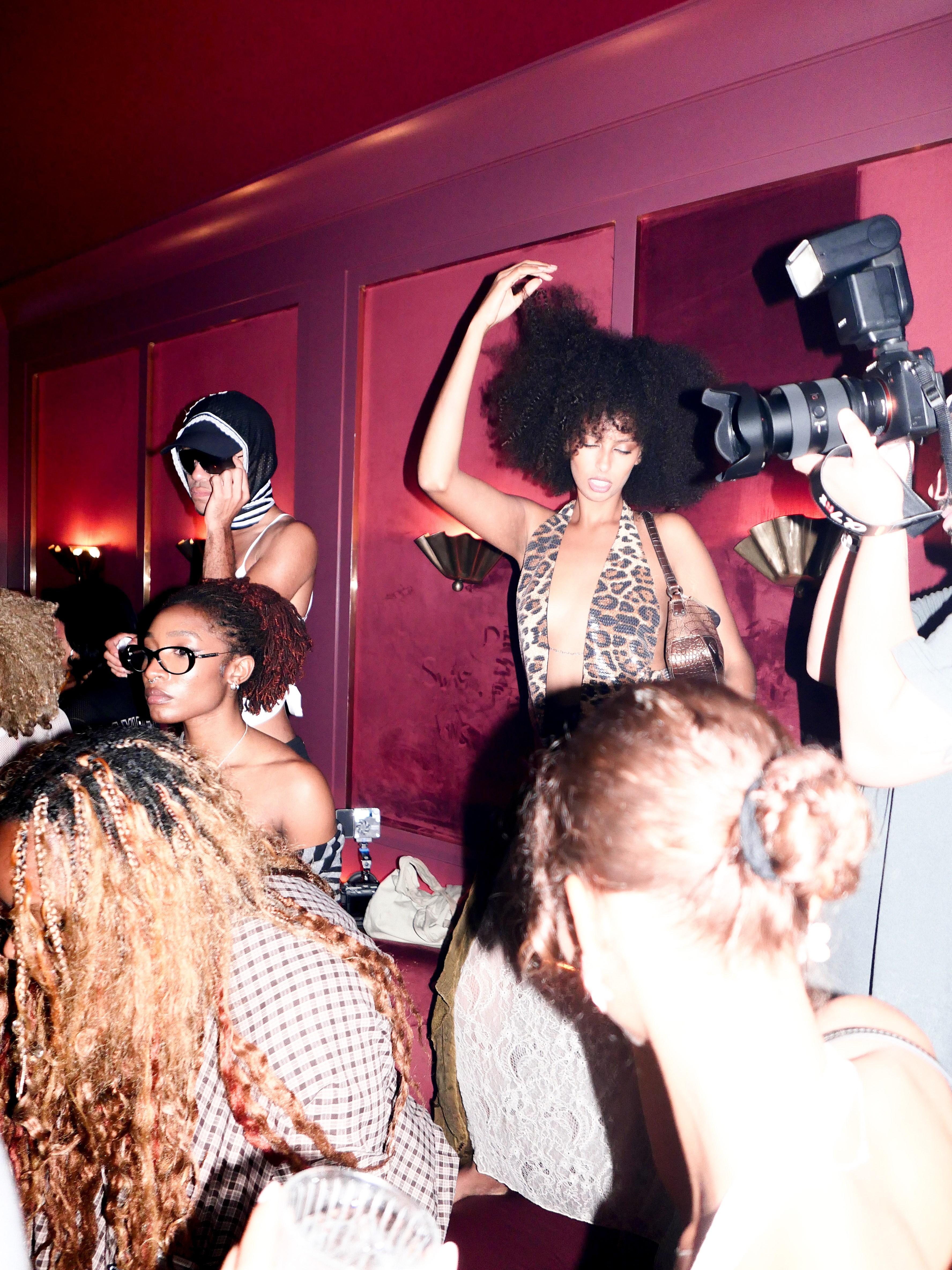 A woman in a leopard print top dances while a photographer captures the scene in a dimly lit, red-walled room.