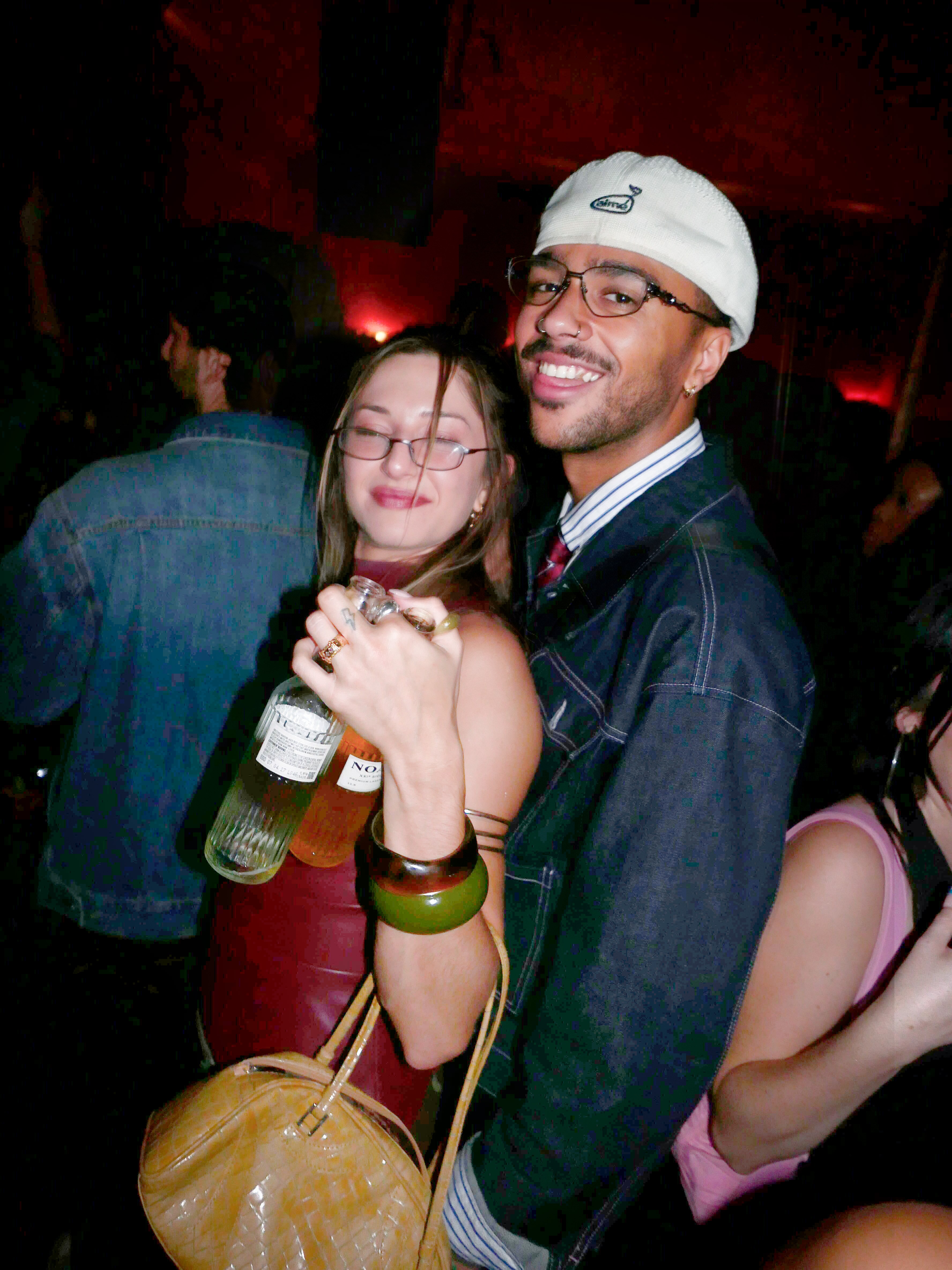 A smiling man in a white beanie and glasses embraces a woman holding two bottles in a dimly lit club.