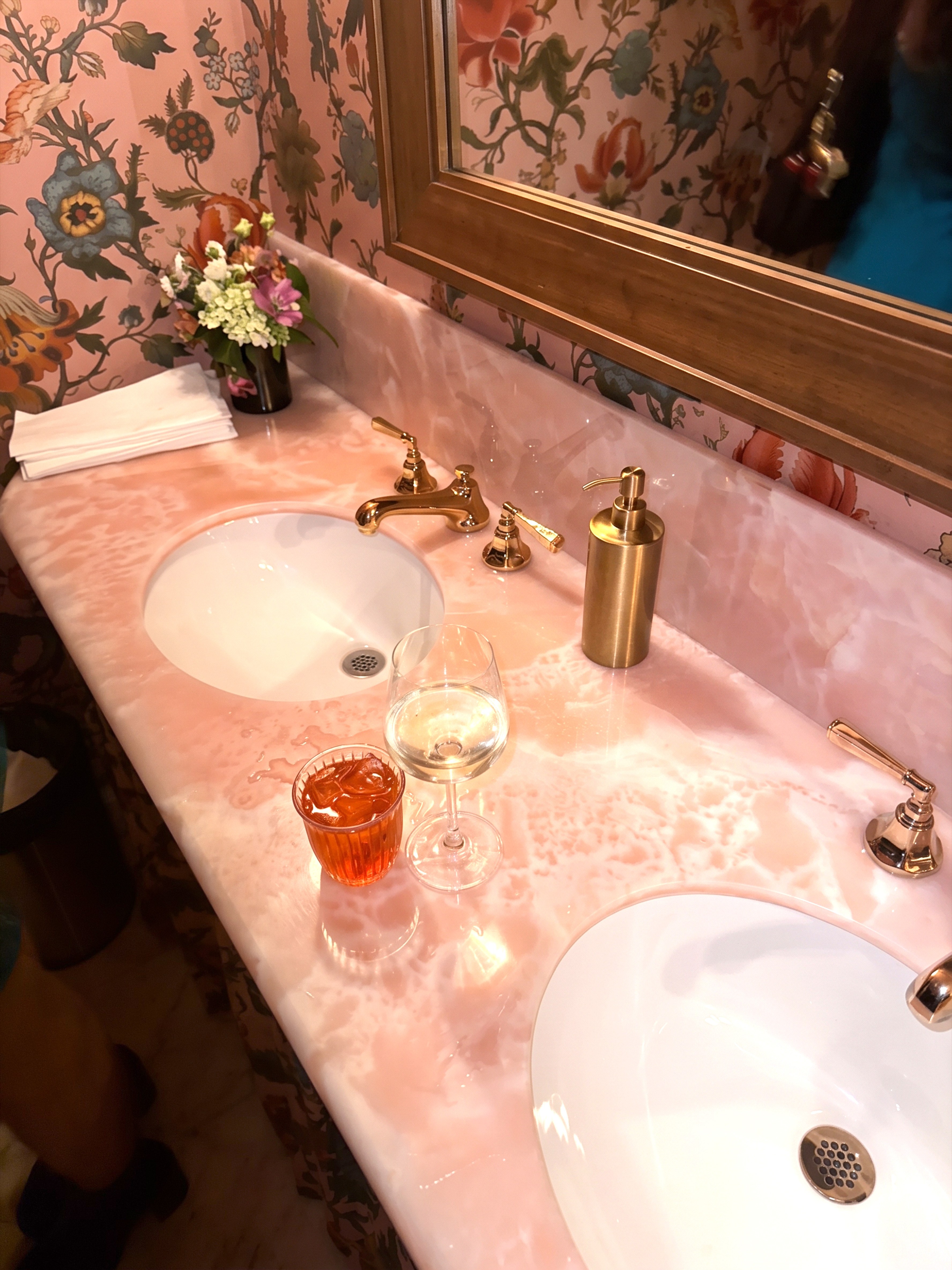 A pink marble vanity with two sinks, gold faucets, a vase of flowers, and two drinks, against floral wallpaper.