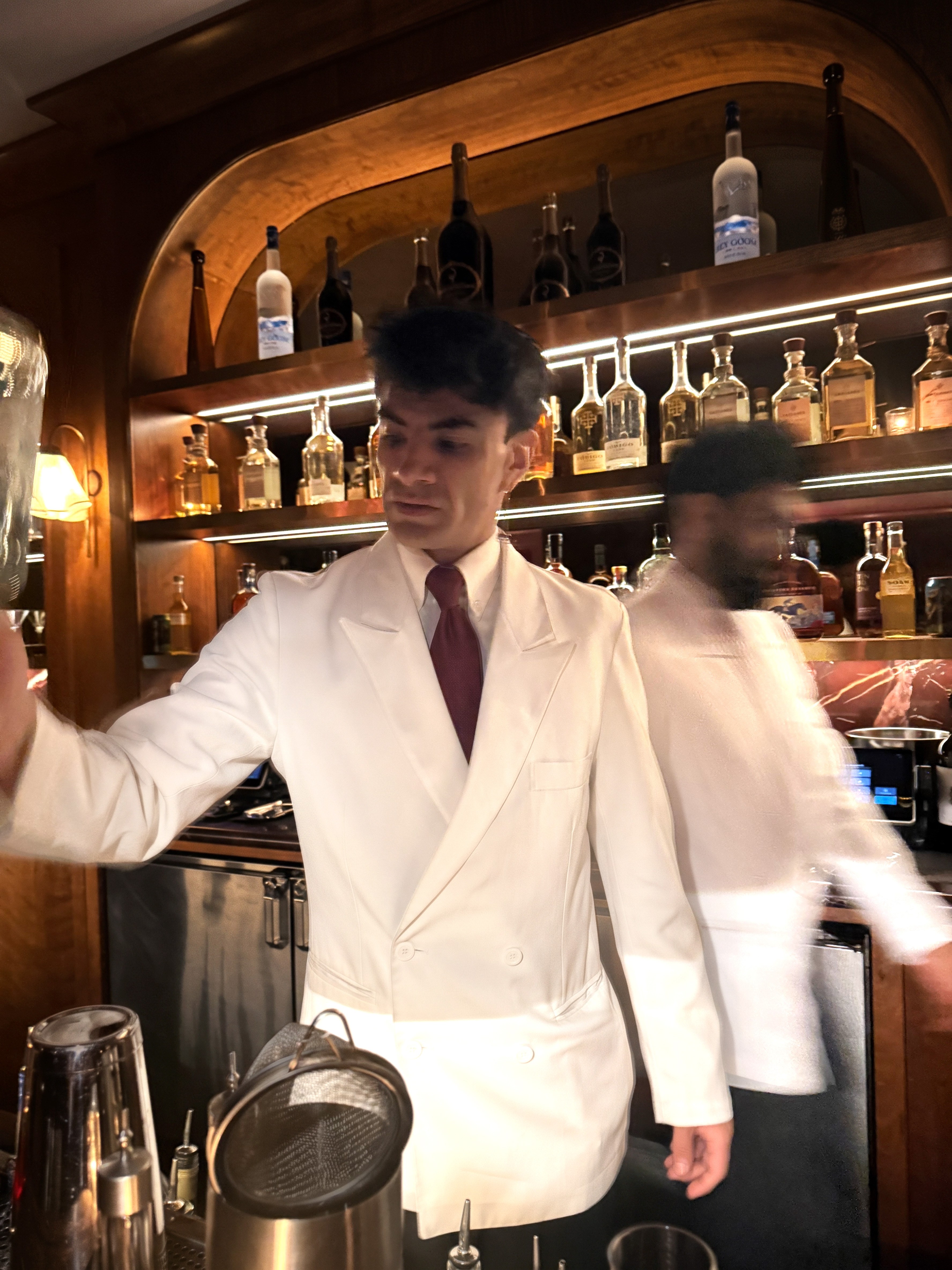 A bartender in a white jacket and red tie mixes a drink in a dimly lit bar with shelves of liquor bottles.