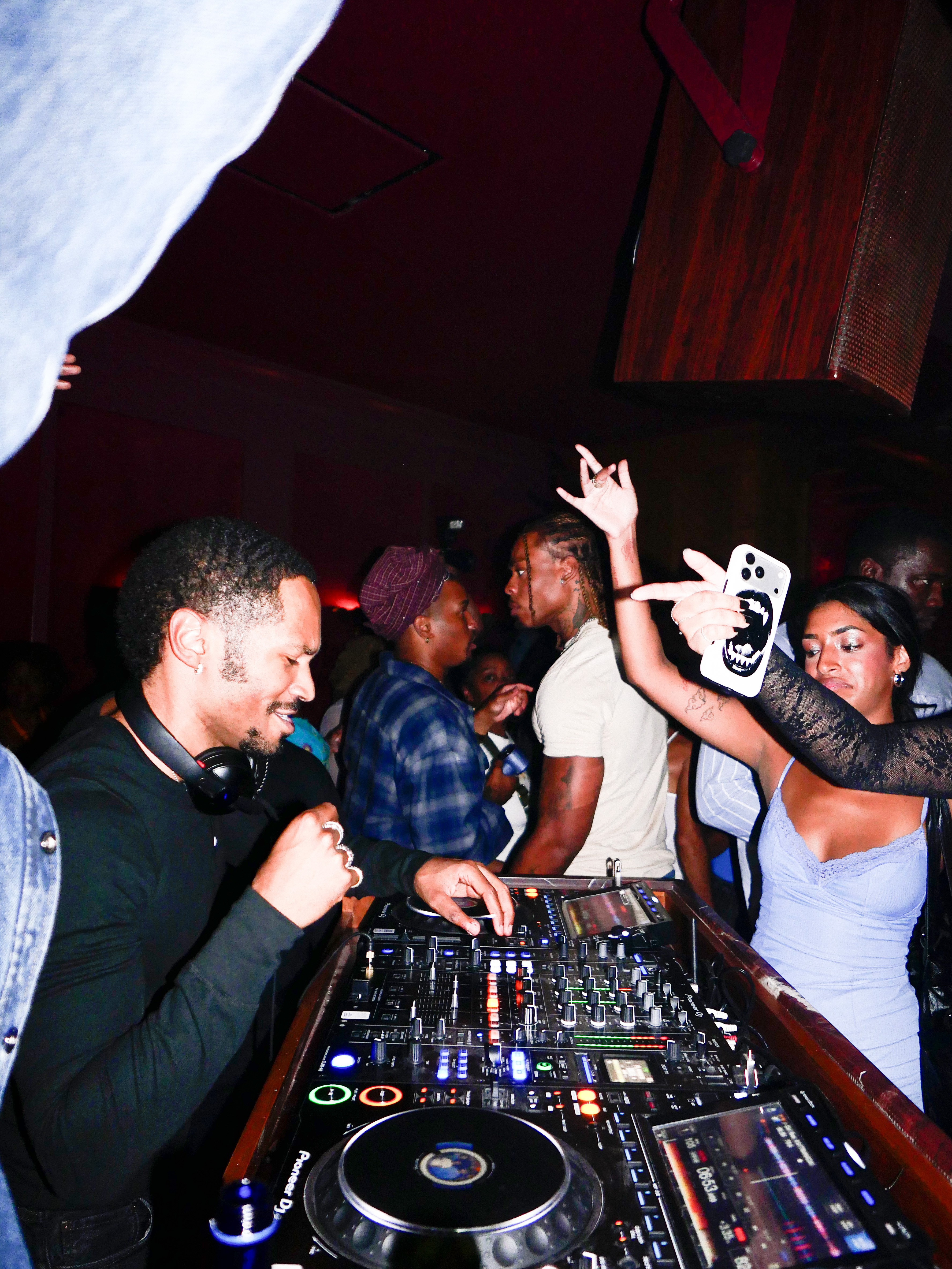 A DJ with headphones around his neck smiles while mixing music on a Pioneer DJ controller, surrounded by dancing club-goers.