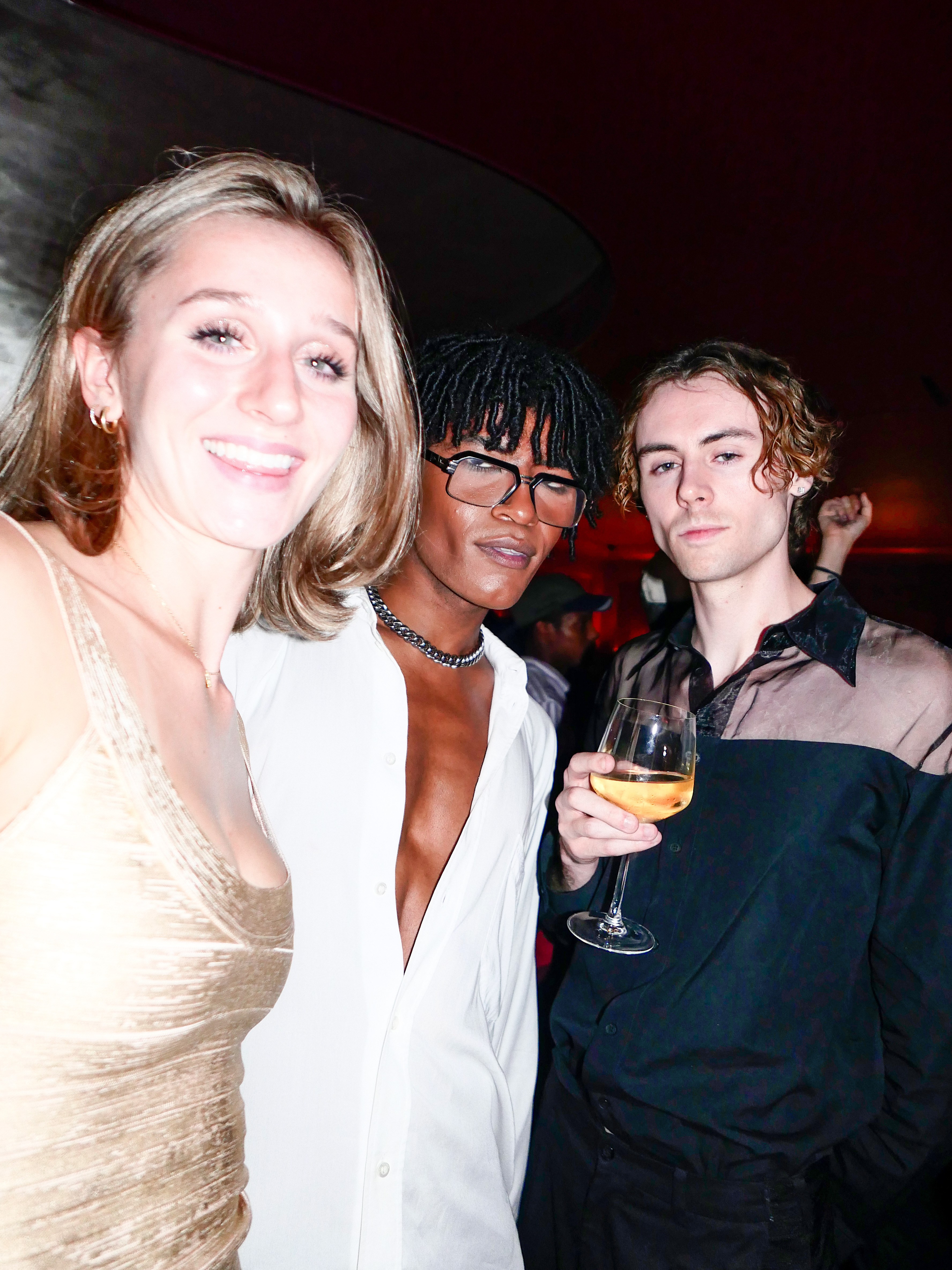 Three young adults at a party: a blonde woman smiling, a Black man in glasses, and a white man holding a wine glass.