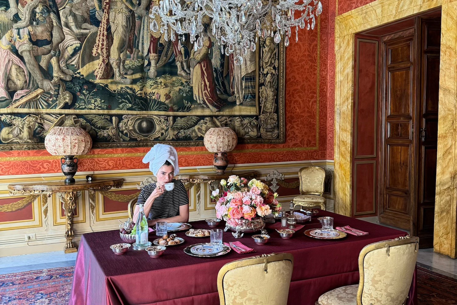 Woman in a towel turban sips from a teacup at a lavishly set table in a room with tapestries and a chandelier.