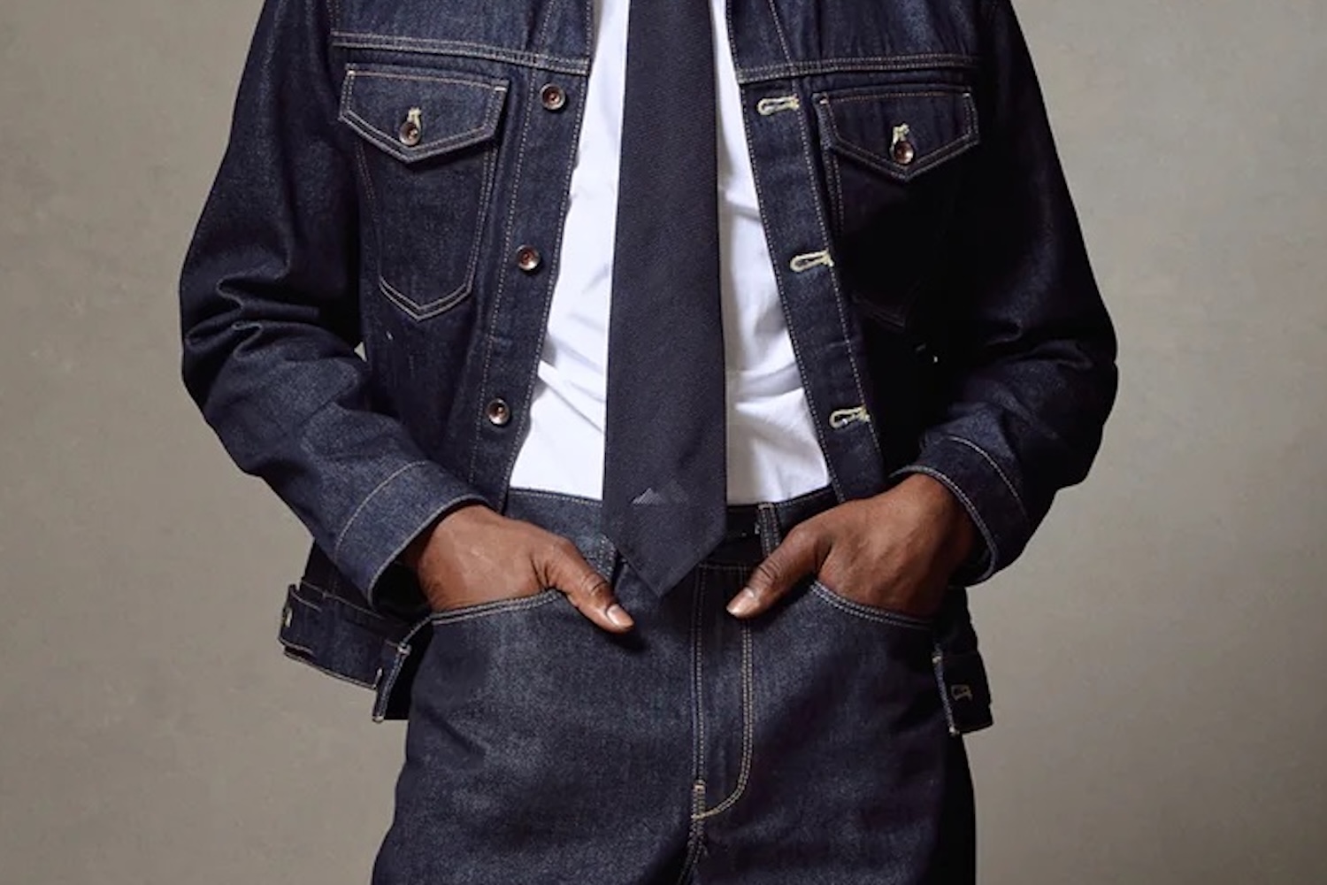 Torso of a person wearing a dark denim jacket and pants, white shirt, and dark tie, hands in pockets.