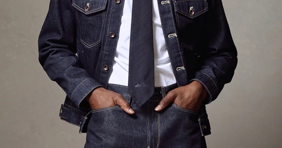 Torso of a person wearing a dark denim jacket and pants, white shirt, and dark tie, hands in pockets.