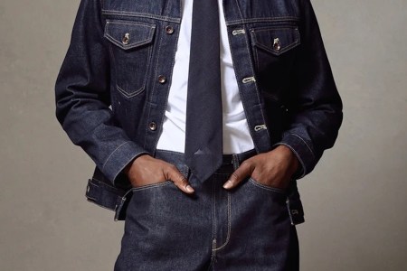 Torso of a person wearing a dark denim jacket and pants, white shirt, and dark tie, hands in pockets.