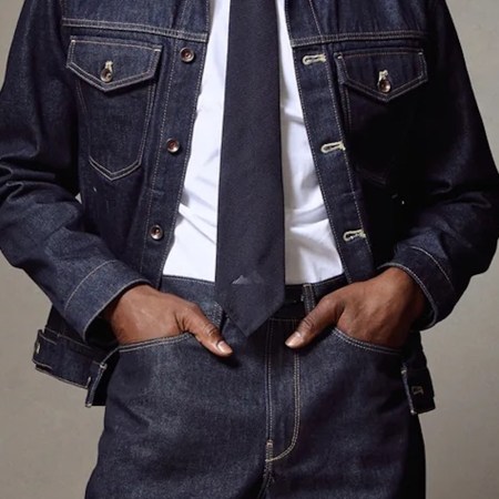 Torso of a person wearing a dark denim jacket and pants, white shirt, and dark tie, hands in pockets.