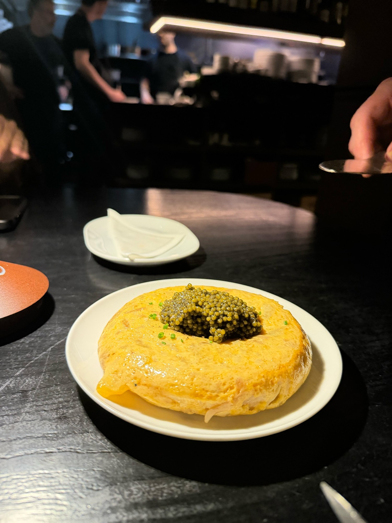 Golden Spanish tortilla topped with a mound of dark caviar and chives on a white plate in a dimly lit restaurant.