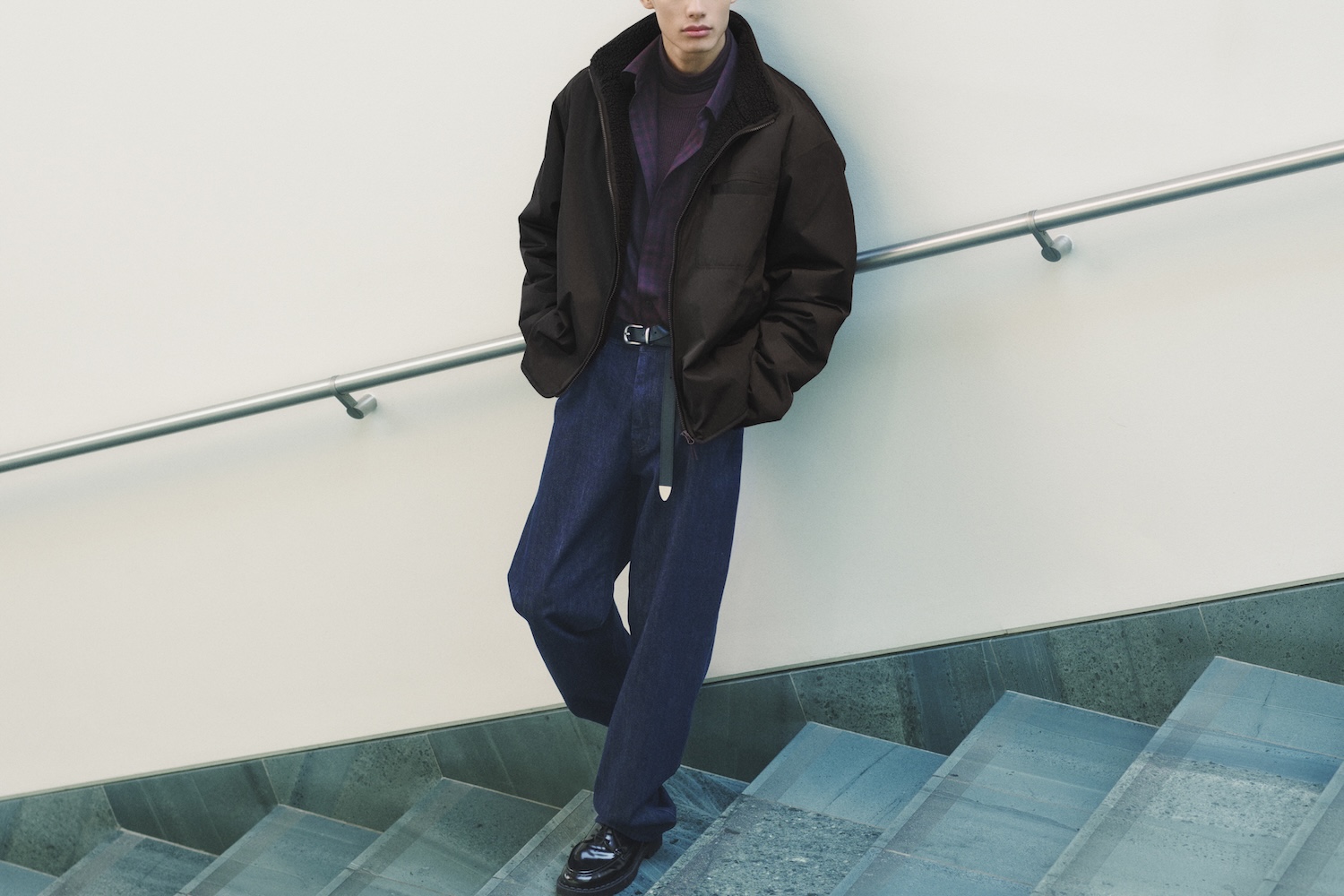 Man in a black puffer jacket with a sherpa collar, purple turtleneck, dark blue jeans, and black loafers on a staircase.