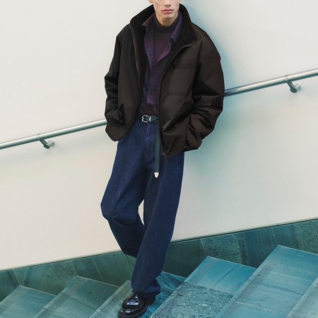 Man in a black puffer jacket with a sherpa collar, purple turtleneck, dark blue jeans, and black loafers on a staircase.