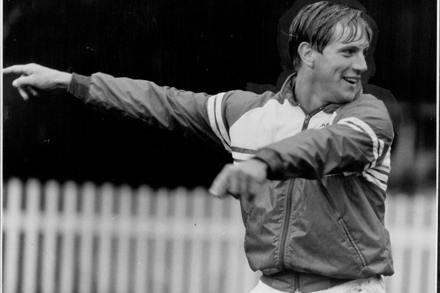 Smiling man in a track jacket, with short hair, points his left arm to the left.