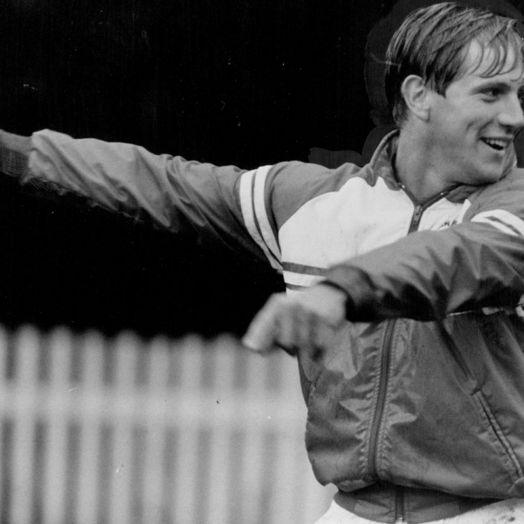 Smiling man in a track jacket, with short hair, points his left arm to the left.