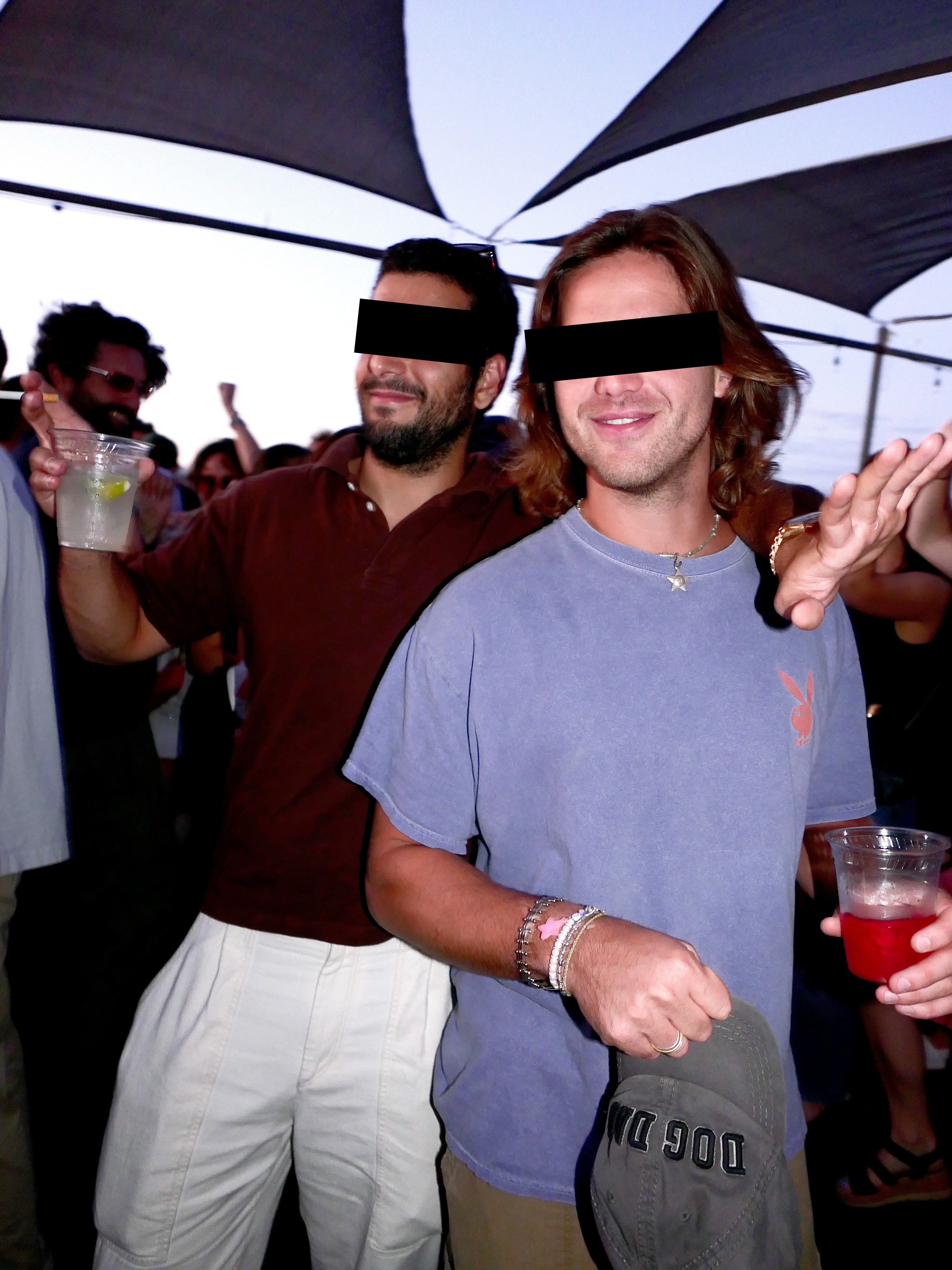 Mikey (left) and George (right) with black bars over their eyes, one in a maroon polo and white pants, the other in a blue t-shirt with a Playboy bunny logo, holding drinks at Toy Tonics party.