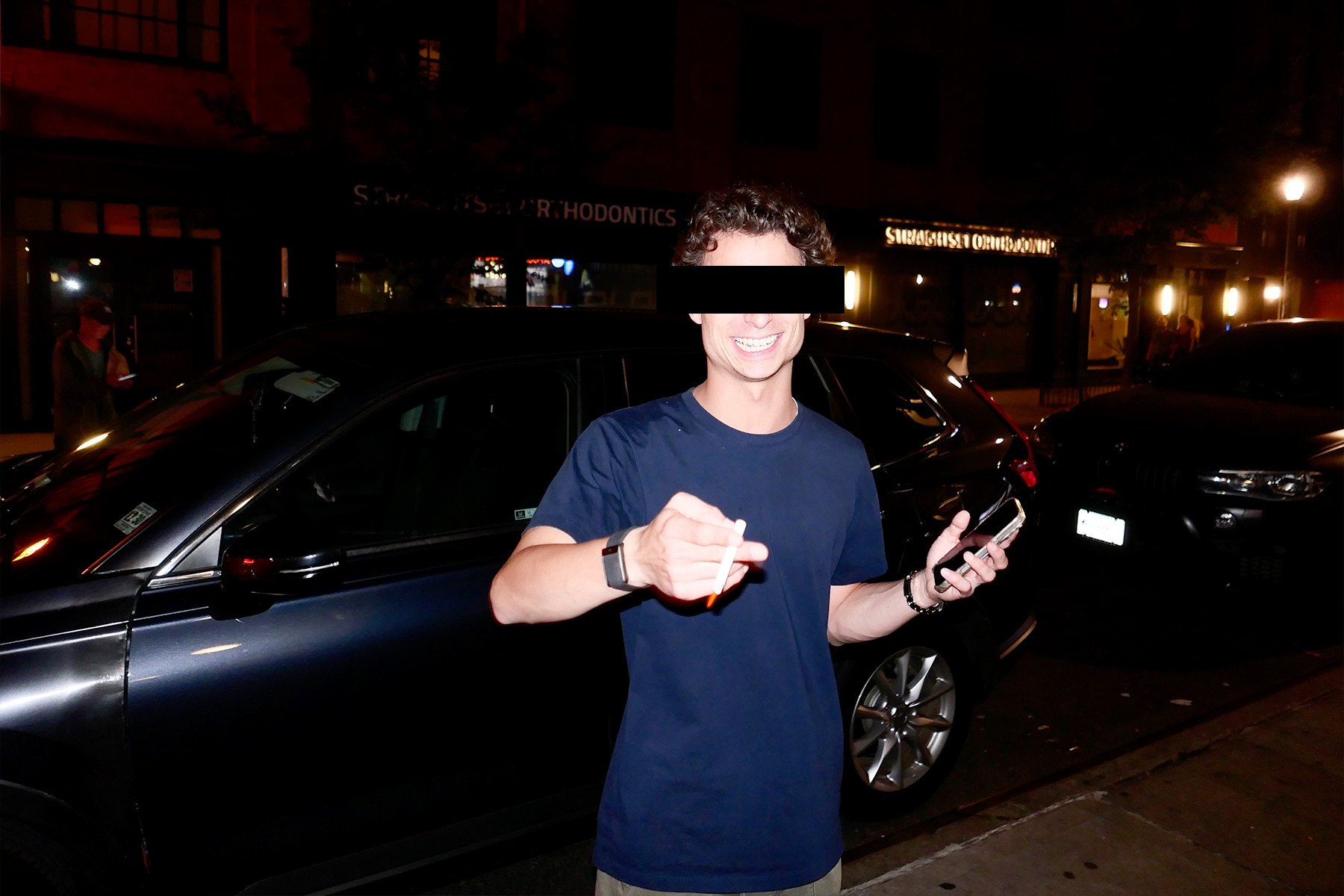 Smiling man (Edu) in a navy t-shirt holding a vape and phone, standing on a street at night with parked cars.