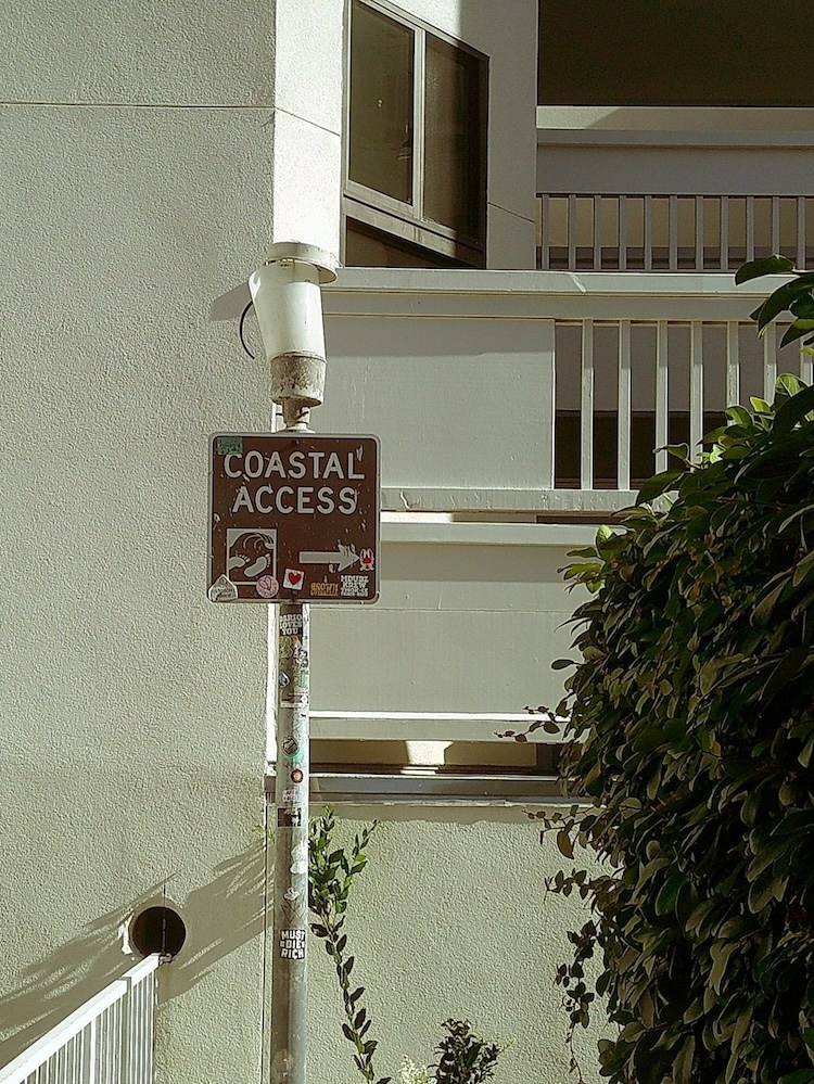 A "Coastal Access" sign with stickers on a pole, next to a building with balconies and lush green foliage.
