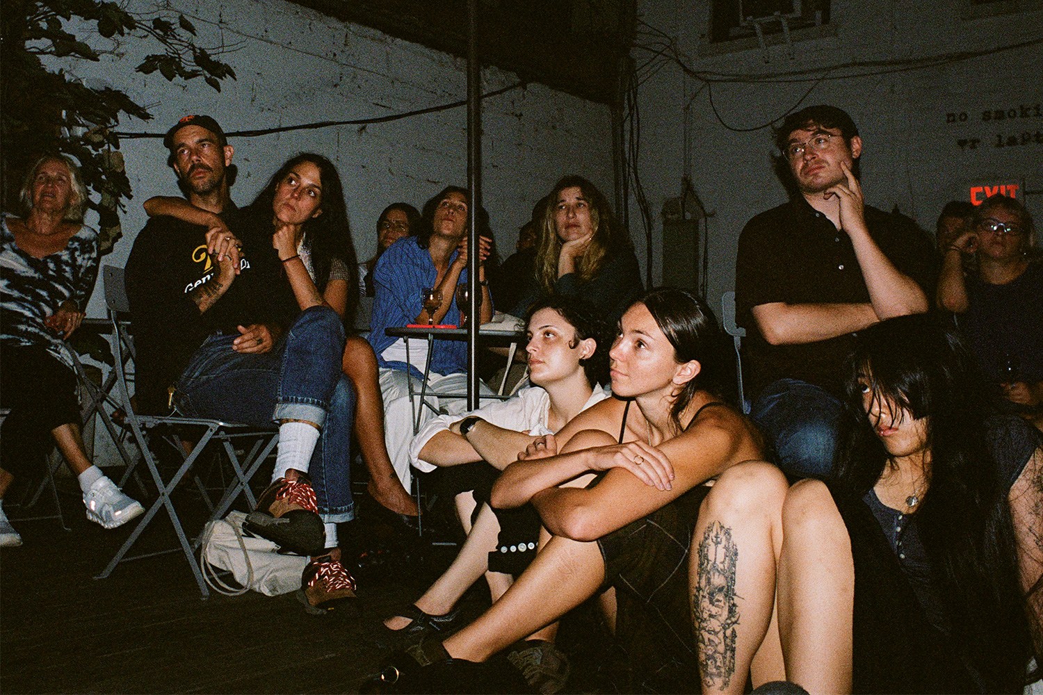 A diverse group of people, some seated, some on the floor, intently watching something off-camera in a dimly lit room.