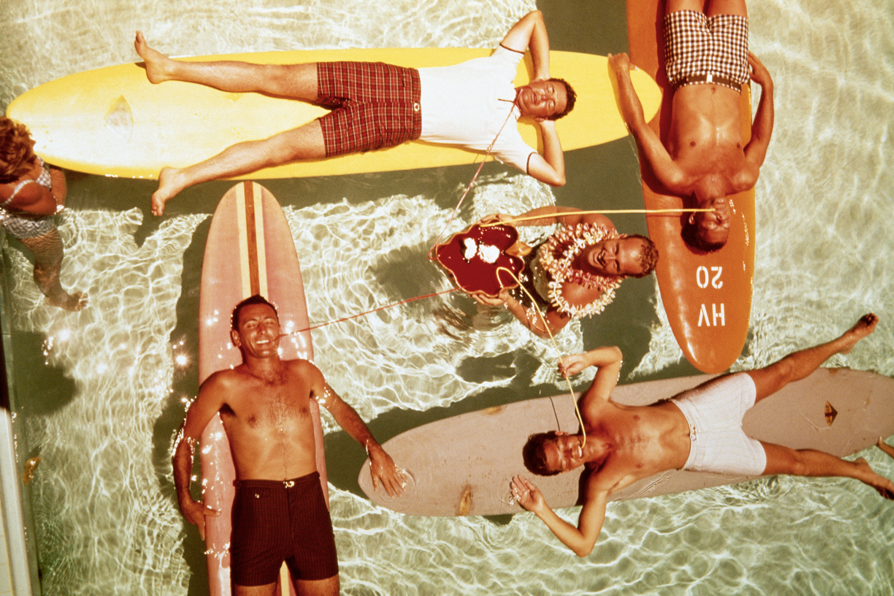 Overhead view of five men on surfboards in a pool, some drinking from straws, one wearing a lei.