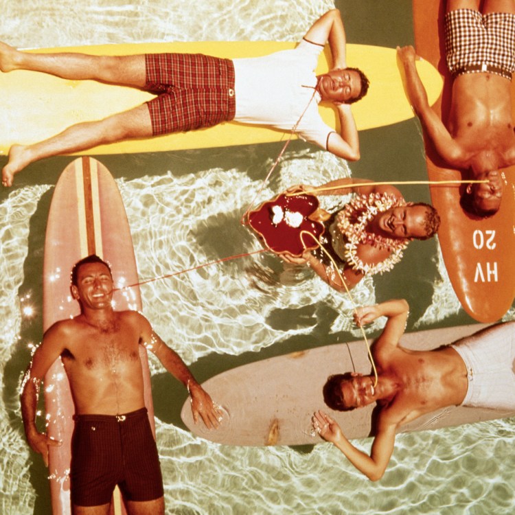 Overhead view of five men on surfboards in a pool, some drinking from straws, one wearing a lei.