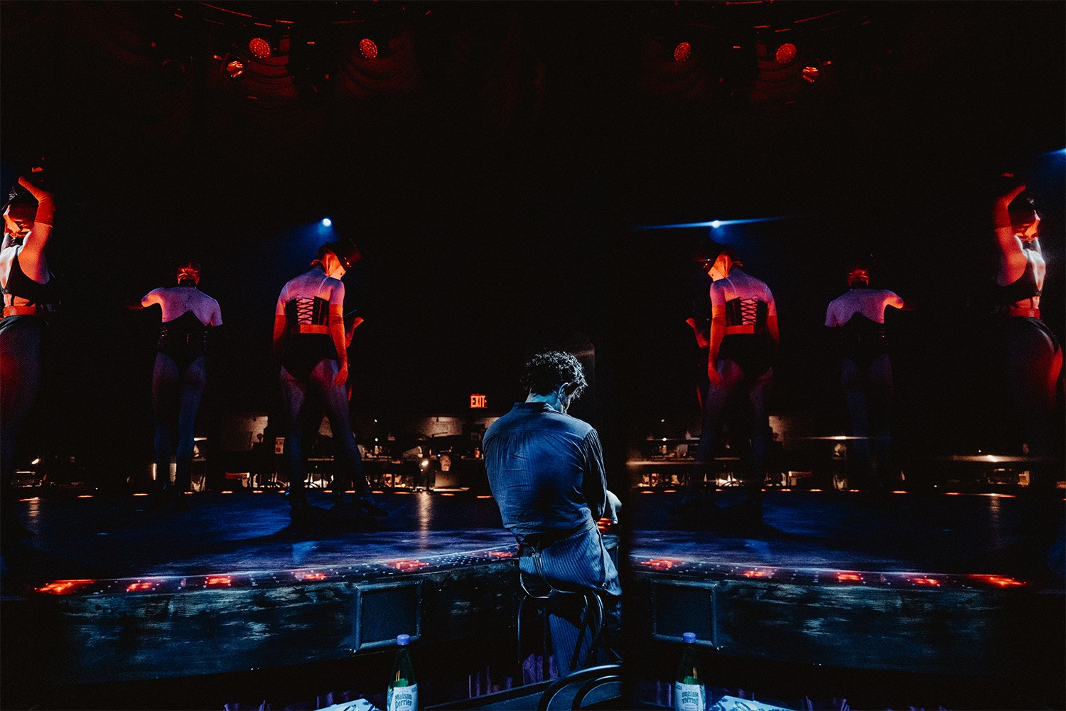 A man stands with his back to us while women in lingerie lit in red light stand on a stage above him