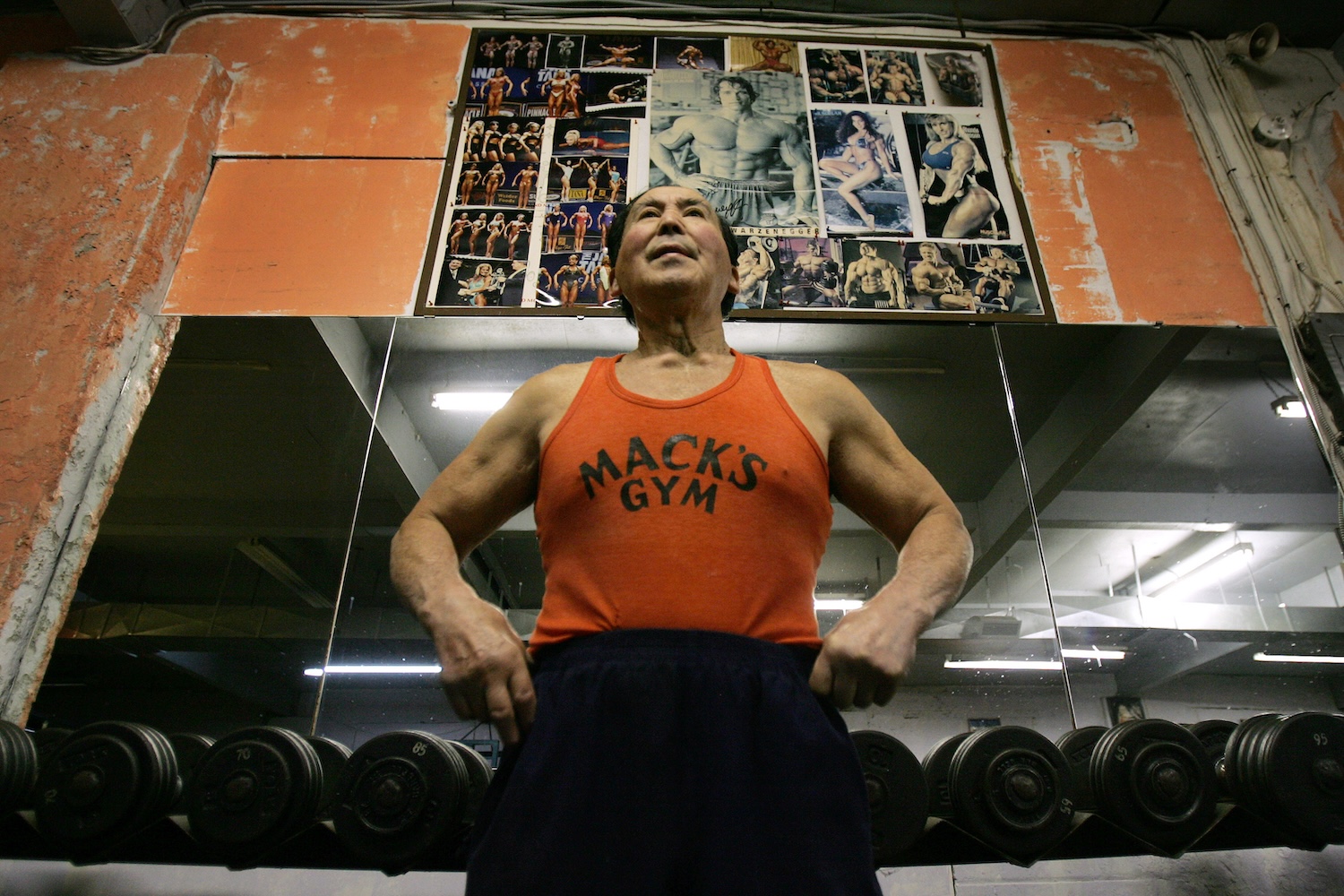 A muscular older man in an orange "Mack's Gym" tank top stands in a gym, with bodybuilding posters above him.